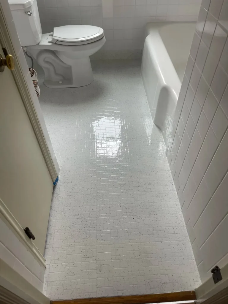 A view down a small bathroom hallway showing a white toilet on the left and a white bathtub entrance on the right. The floor is covered in glossy white mosaic tile with small dark flecks.