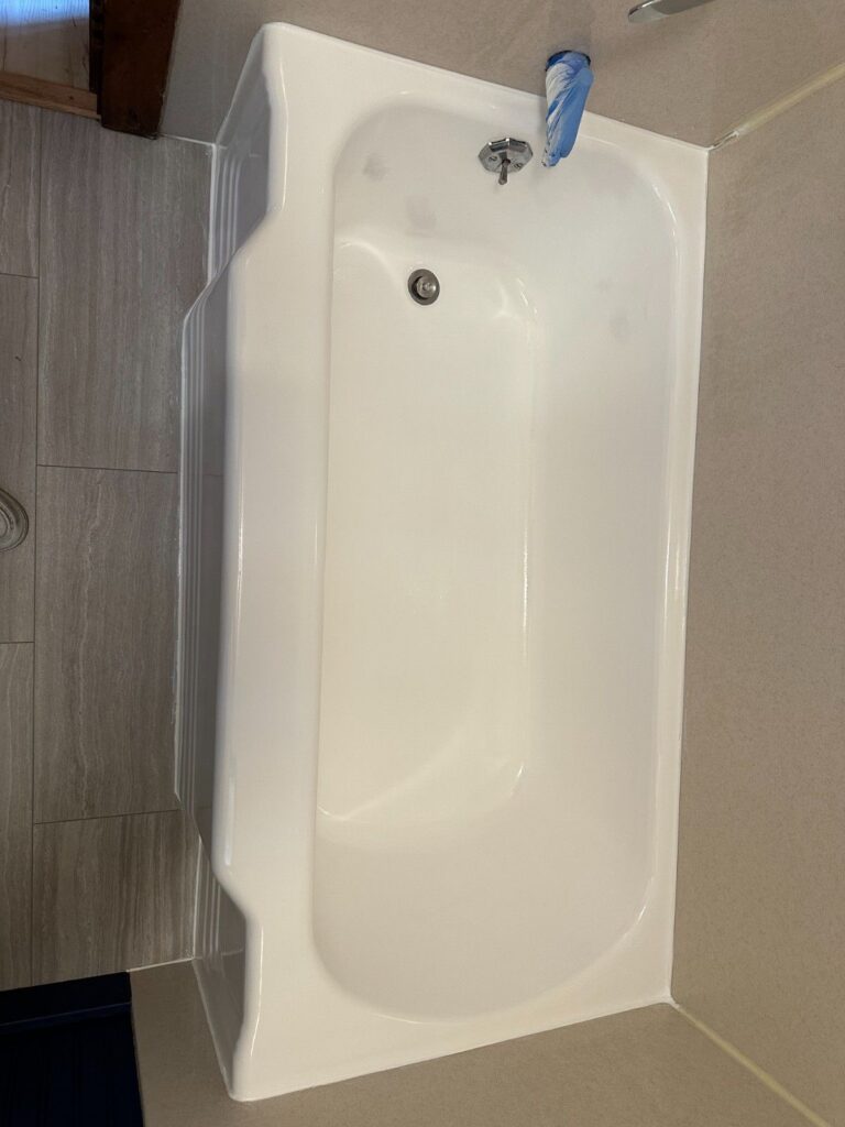 Top-down view of a shiny white bathtub with modern, light gray tile on the surrounding floor and wall. A blue scrubber brush is visible near the faucet.