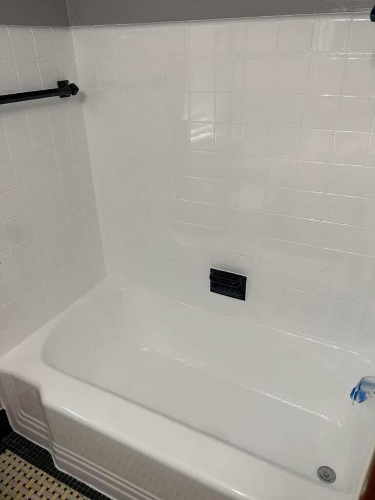 Close-up view of a white bathtub and surrounding white tiled walls, which appear to have been recently refinished. There is a black towel bar on the left wall and a black soap dish holder inside the tub area.