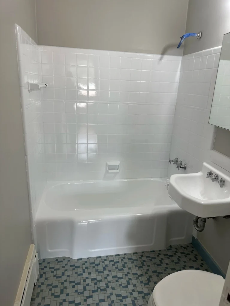 Full view of a bathroom showing a white bathtub, white tiled walls, a wall-mounted sink, and a toilet. The floor has a blue and white square mosaic tile pattern. The walls are painted gray.